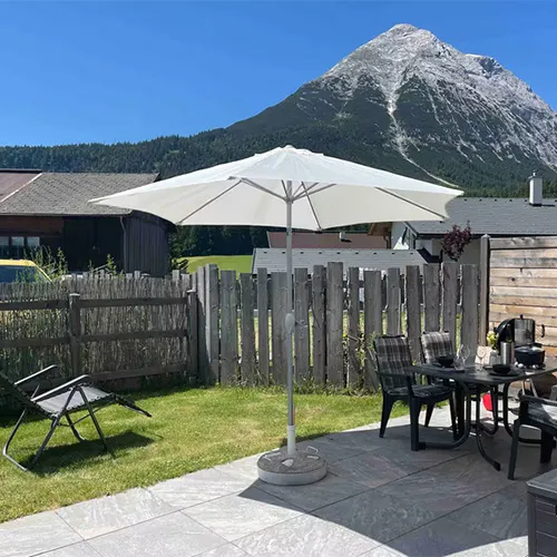 Terrasse mit weißem Sonnenschirm, Sitzmöbeln im Außenbereich und Liegestuhl vor Bergkulisse und blauem Himmel.