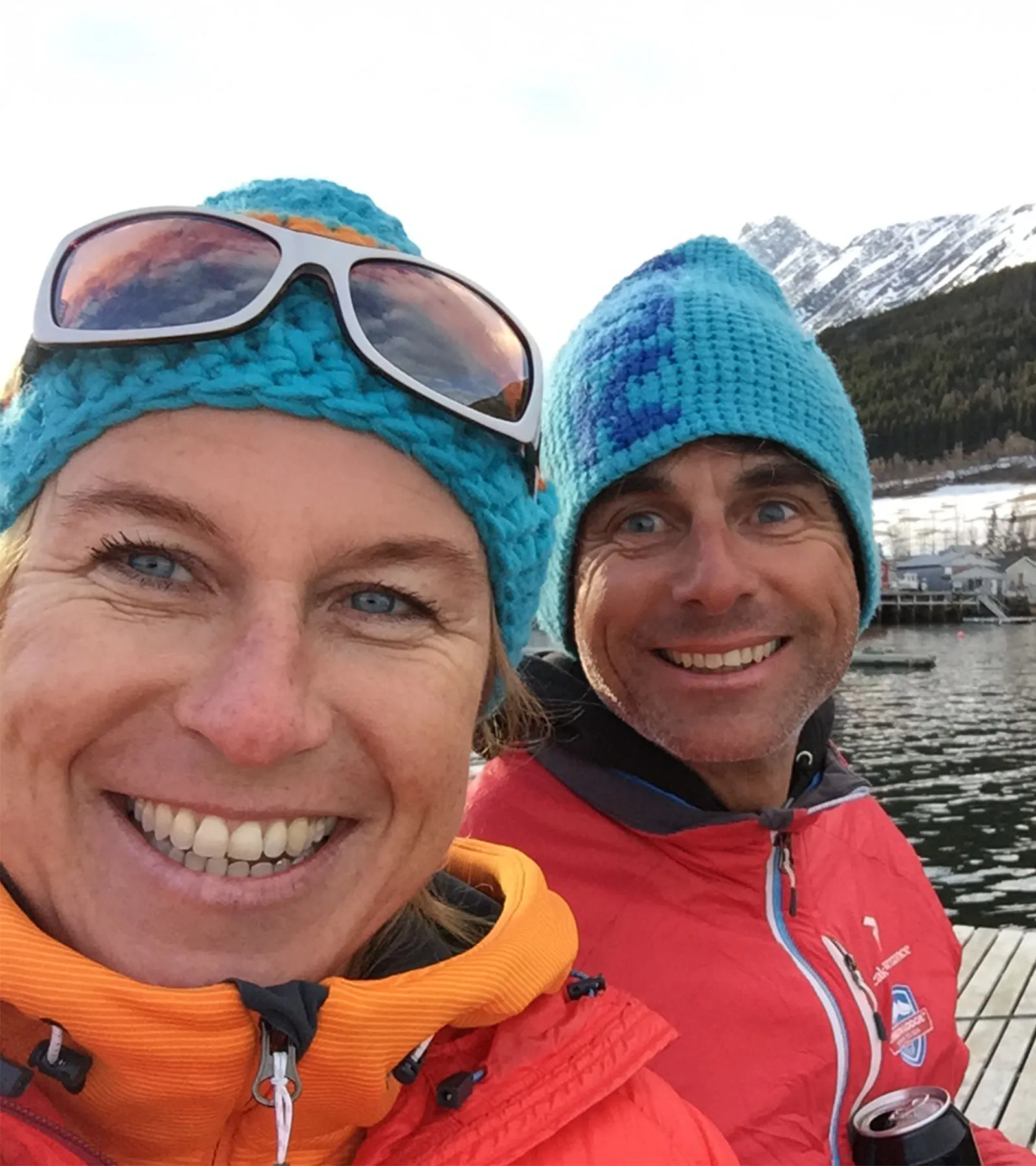 Silke und Burkhard lächeln in bunten Winterjacken und blauen Strickmützen und machen ein Selfie an einem verschneiten Bergsee.