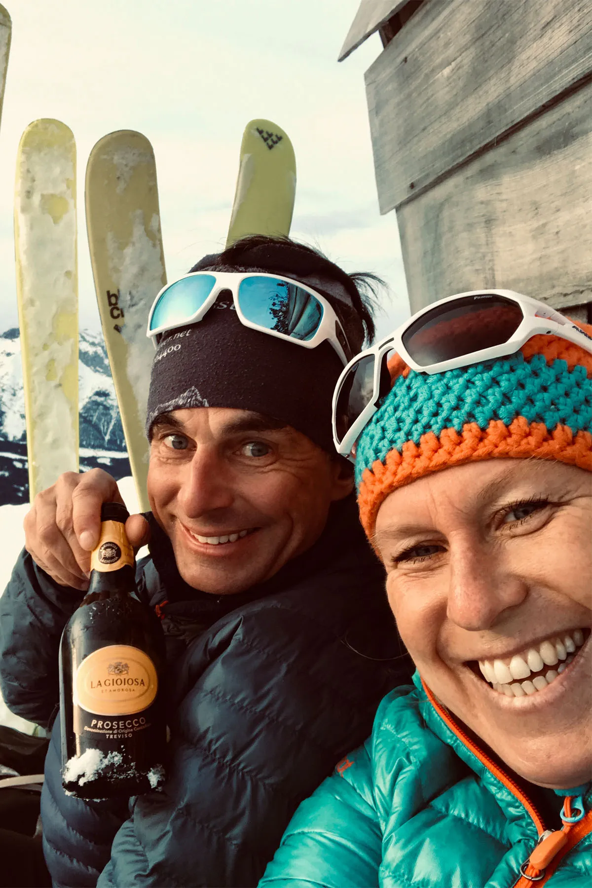Silke und Burkhard lächeln in Winterkleidung und halten eine Flasche La Gioiosa Prosecco, im Hintergrund Skier und verschneite Berge.