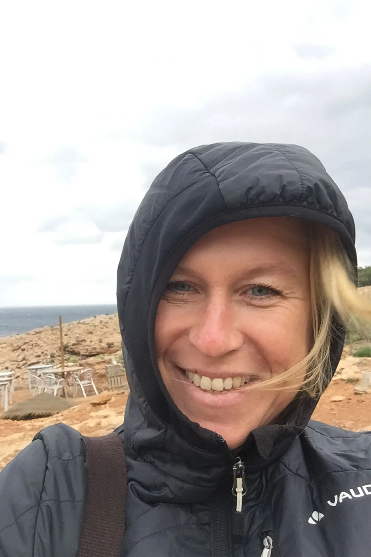 Silke lächelt in schwarzer Kapuzenjacke vor felsiger Landschaft mit Meer im Hintergrund.