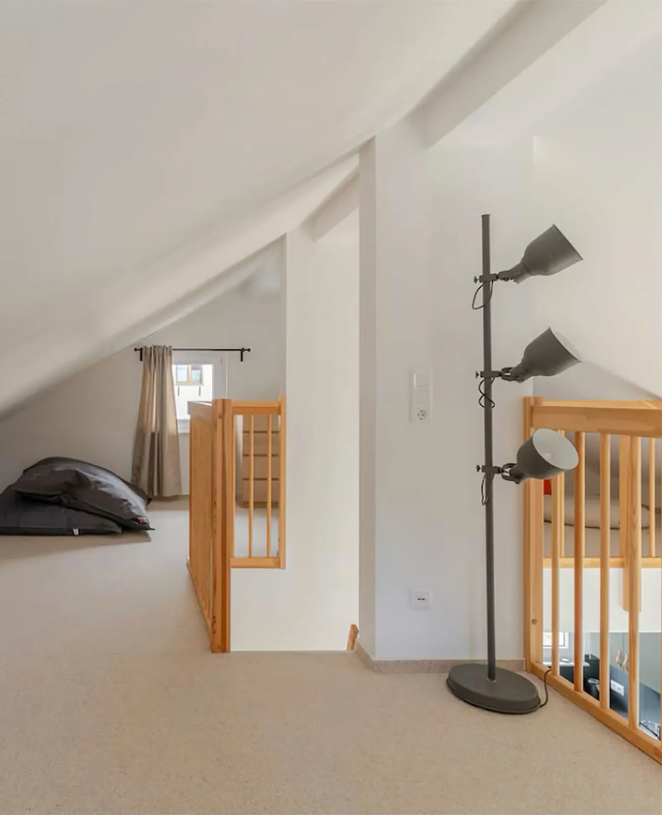 Minimalistisches Obergeschoss mit schräger Decke, Holzgeländer, grauer Stehlampe und Sitzsäcken am Fenster mit beigen Vorhängen.