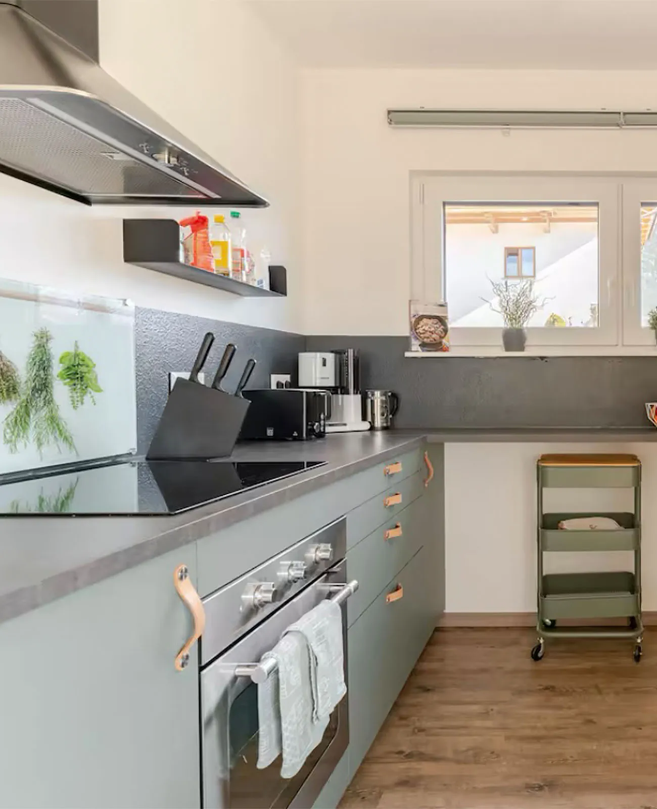 Moderne Küche mit hellgrauen Schränken, Edelstahl-Backofen und Kochfeld, Küchenutensilien und Fenster über der Arbeitsfläche.