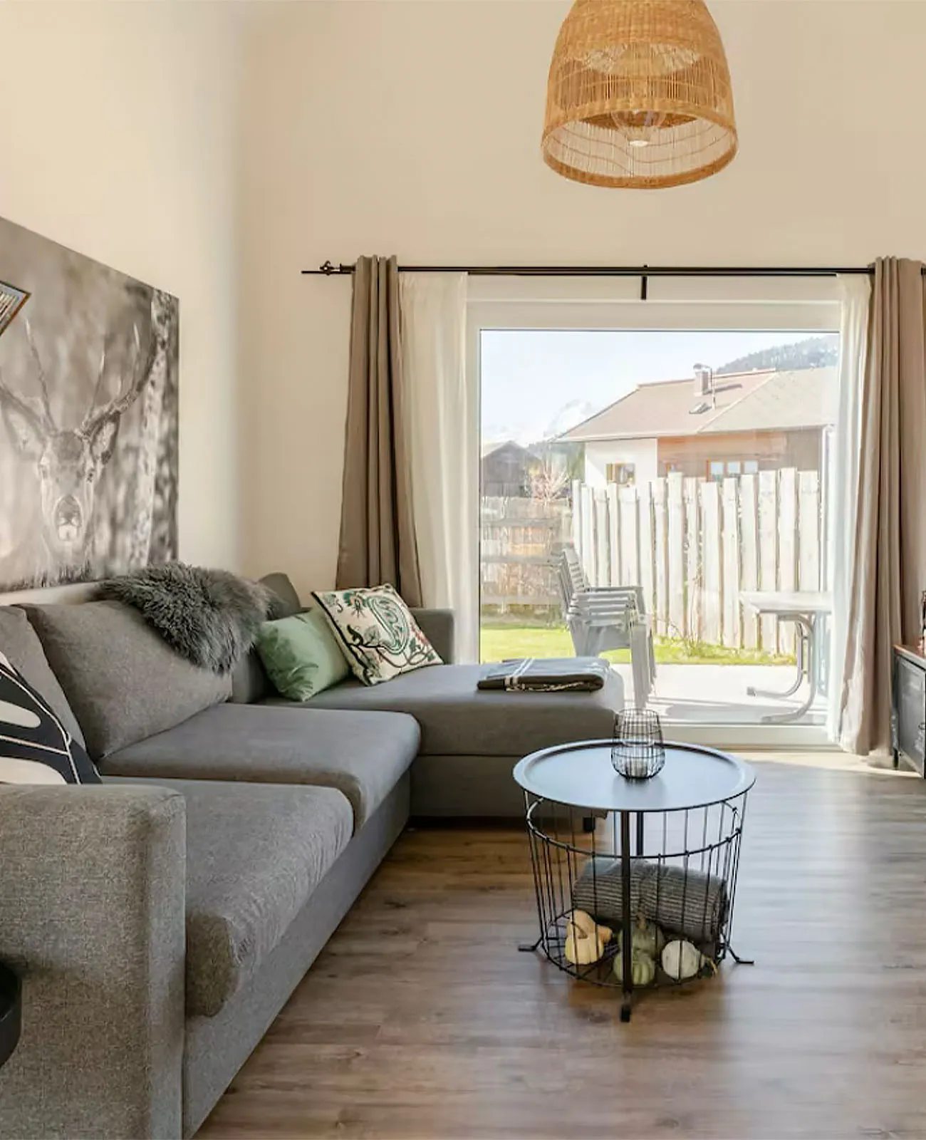 Wohnzimmer mit grauem Ecksofa, dekorativen Kissen, rundem Draht-Couchtisch und großem Fenster mit Blick auf Sitzmöbel im Außenbereich und ein Haus.