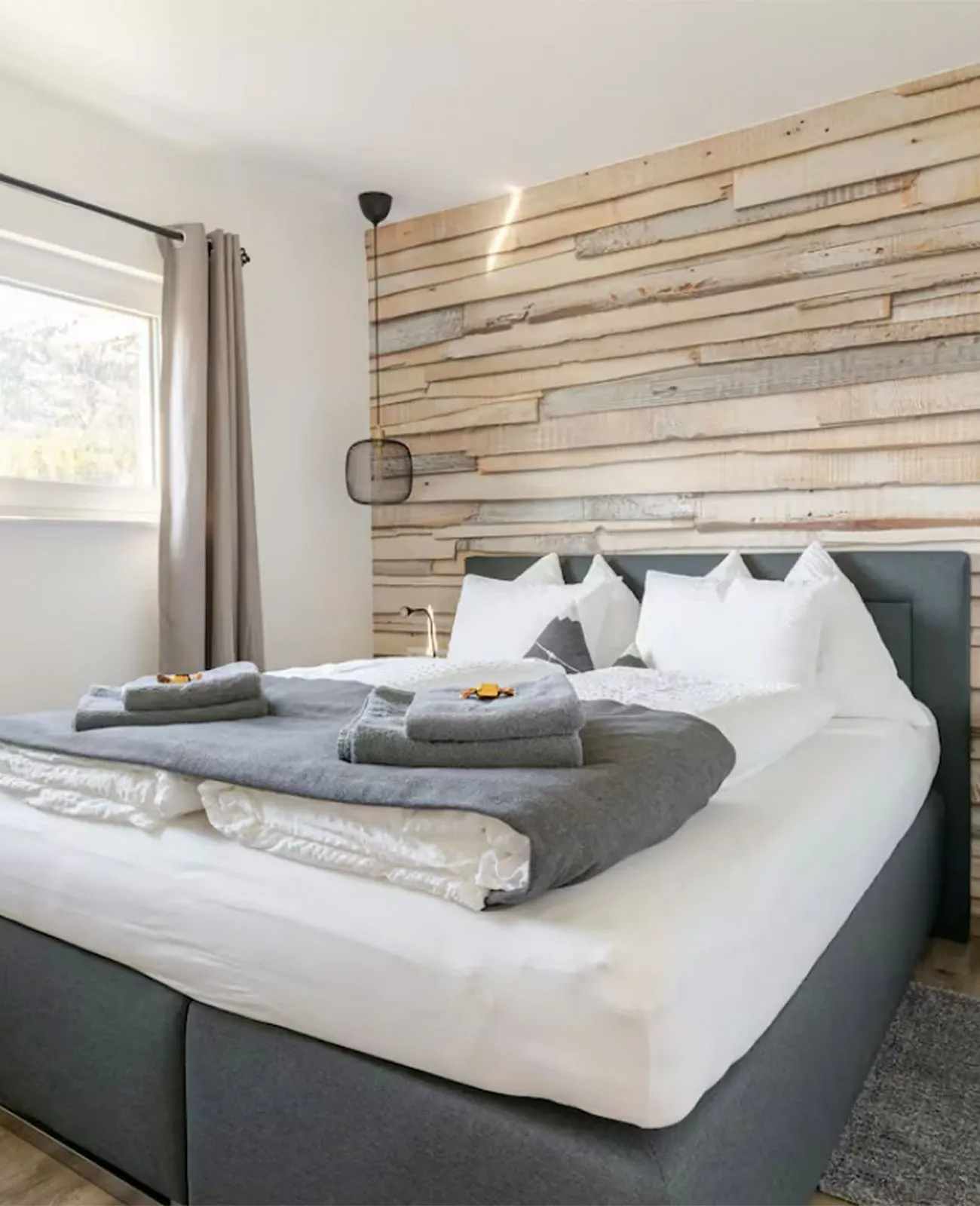 Modernes Schlafzimmer mit großem Bett, weißer Bettwäsche, grauer Decke und gefalteten Handtüchern vor einer Akzentwand aus Holzpaneelen.
