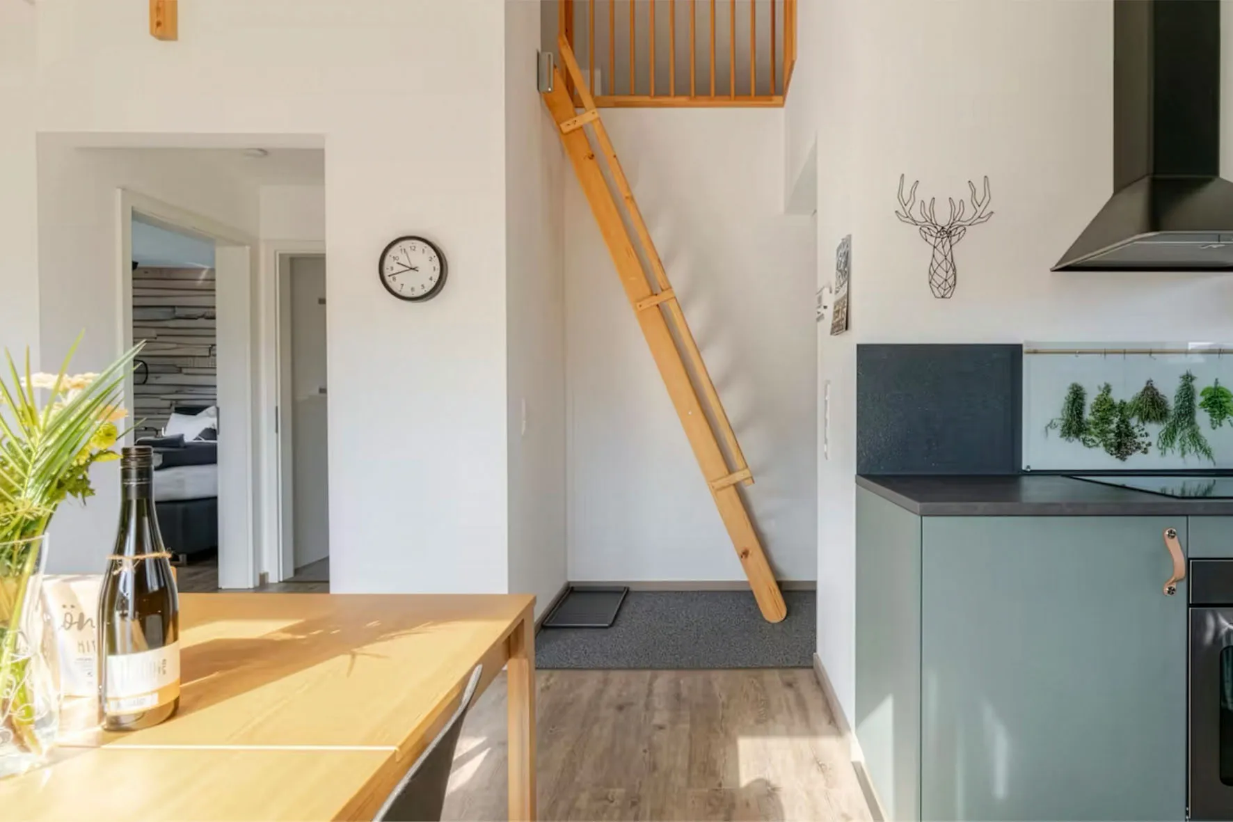 Helle, moderne Küche mit Essbereich, Holztisch, Weinflasche, hängenden Kräutern und Holzleiter zum Obergeschoss.