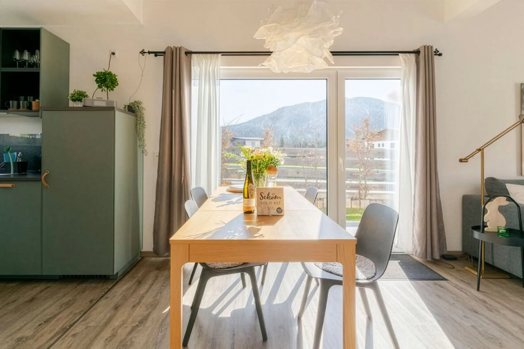 Heller, moderner Essbereich mit Holztisch, grauen Stühlen und großen Glastüren mit Blick auf die Berge.
