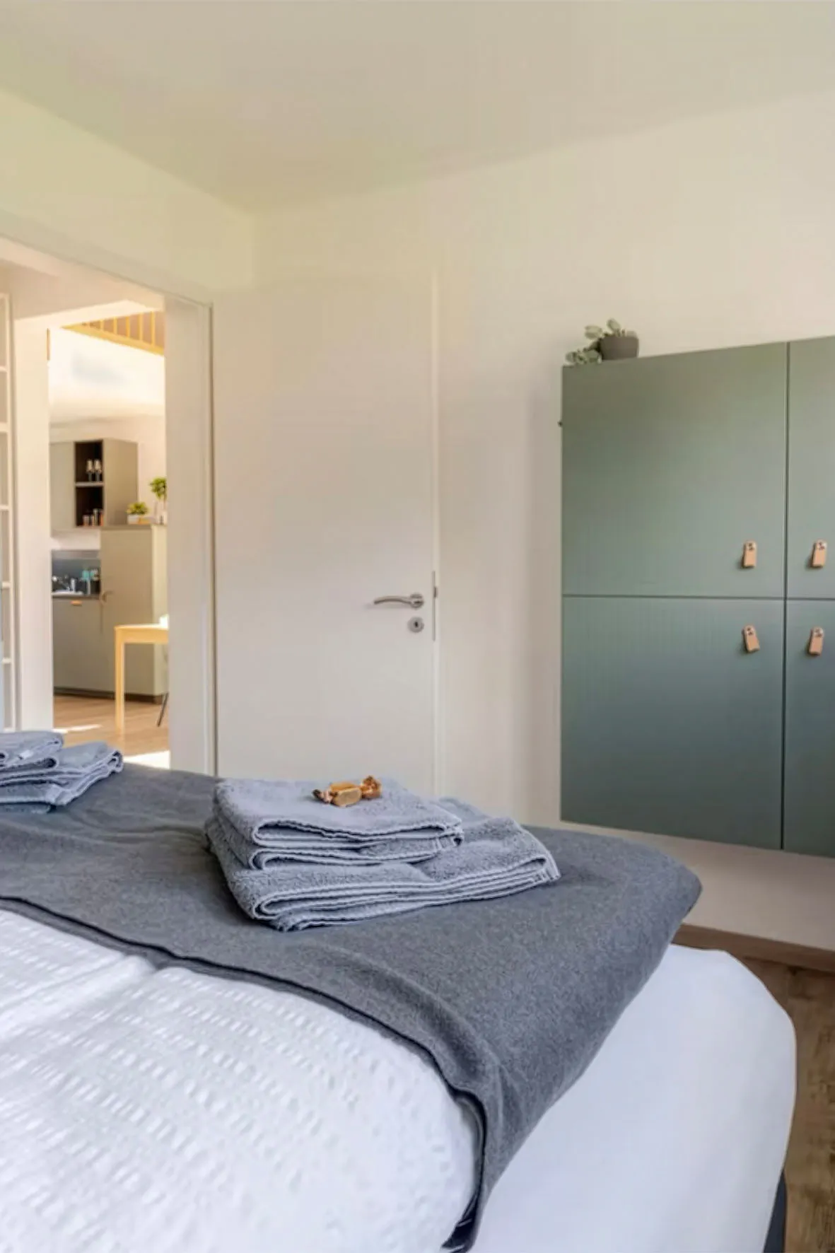Ordentlich gemachtes Bett mit gefalteten grauen Handtüchern, geschlossener weißer Tür und grünen Wandschränken in einem hellen Schlafzimmer.
