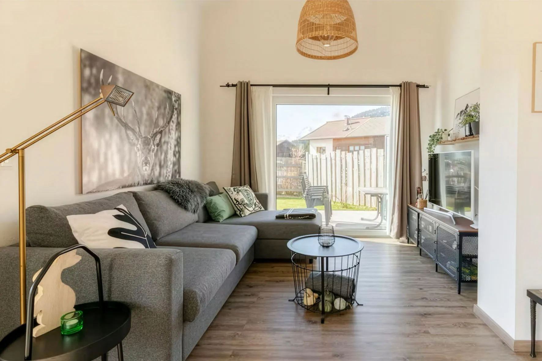 Helles Wohnzimmer mit grauem Ecksofa, dekorativen Kissen, rundem schwarzem Couchtisch und großem Fenster mit Blick in einen eingezäunten Garten.