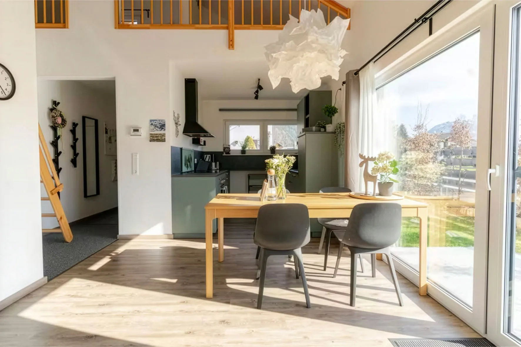 Sonnendurchfluteter, moderner Essbereich mit hellem Holztisch, vier grauen Stühlen, Blumen auf dem Tisch und großen Schiebetüren mit Blick in den Garten.