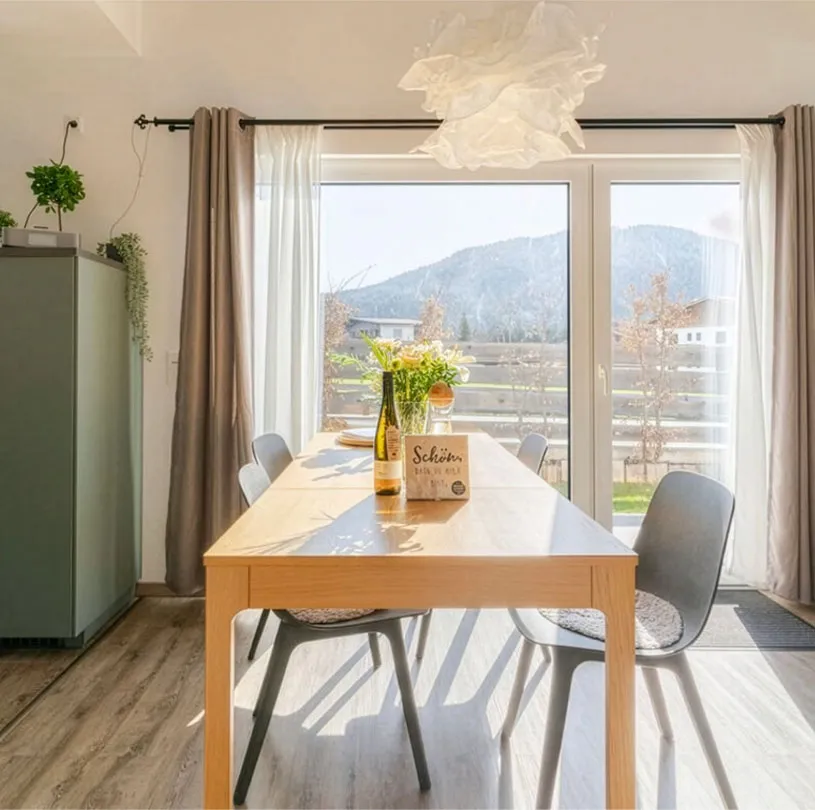 Heller Essbereich mit Holztisch, vier grauen Stühlen, Flasche, Blumenvase und großen Fenstern mit sonnigem Bergblick.