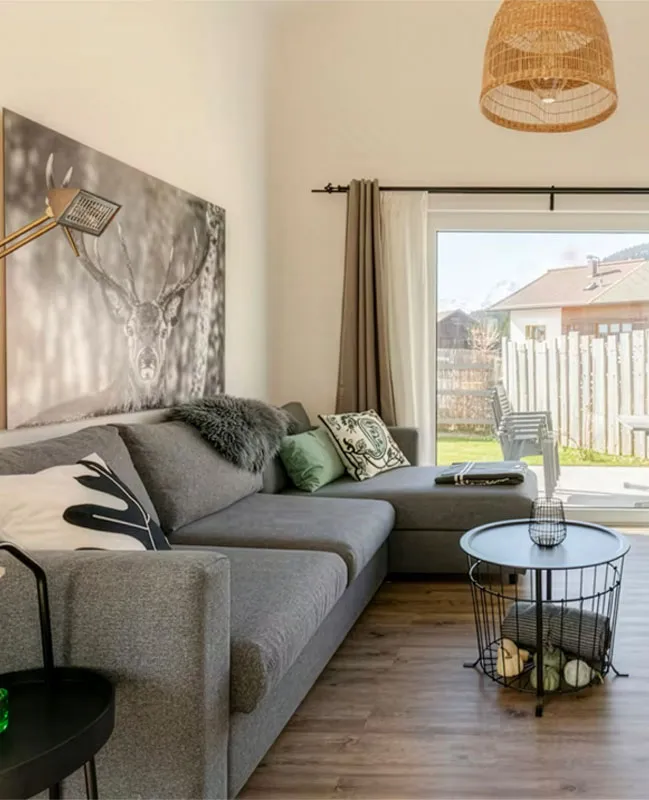 Gemütliches Wohnzimmer mit grauem Ecksofa, dekorativen Kissen, rundem Draht-Couchtisch und großem Fenster mit Blick in einen eingezäunten Garten.