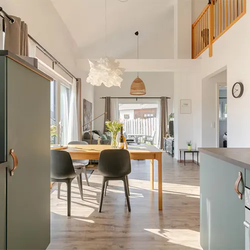 Heller Essbereich mit Holztisch, vier grauen Stühlen, großem Fenster mit Sonneneinfall und moderner Küche im Hintergrund.