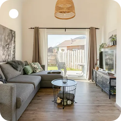 Helles Wohnzimmer mit grauem Ecksofa, rundem Draht-Couchtisch, Schiebetür mit Vorhängen und Fernseher auf schwarzem Lowboard.