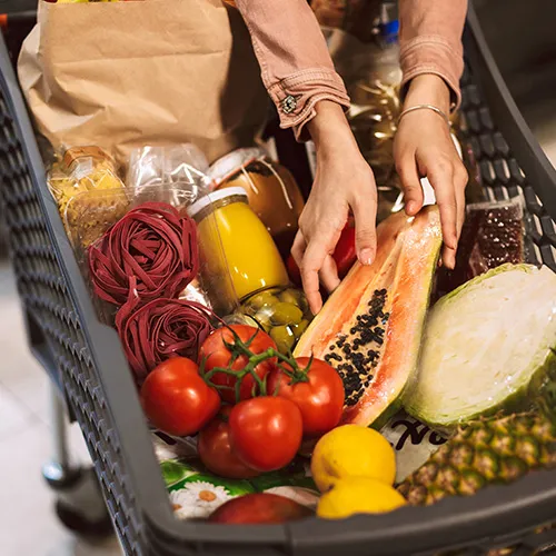 Einkaufswagen voller frischer Tomaten, Zitronen, Kohl, Pasta und Gläser mit Konserven, eine Person ordnet sie.