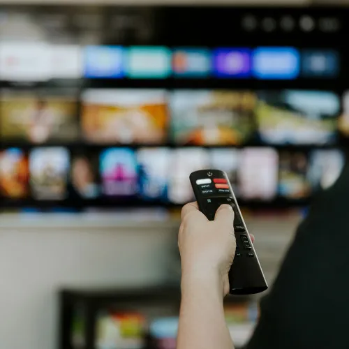 Person hält eine TV-Fernbedienung auf einen unscharfen Bildschirm mit einer Übersicht aus farbigen Film- oder Serienvorschauen gerichtet.