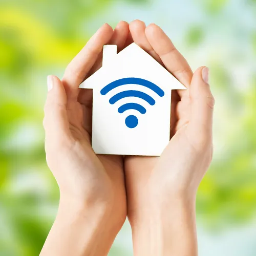 Zwei Hände halten vorsichtig ein weißes Haus-Symbol mit blauem WLAN-Zeichen vor unscharfem Hintergrund.