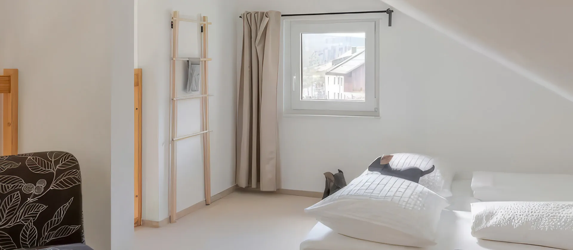 Helles Schlafzimmer mit weißen Wänden, kleinem Fenster mit beigen Vorhängen, Bett mit weißen Kissen und Bettwäsche sowie einem Holzständer für Handtücher oder Kleidung in der Ecke.
