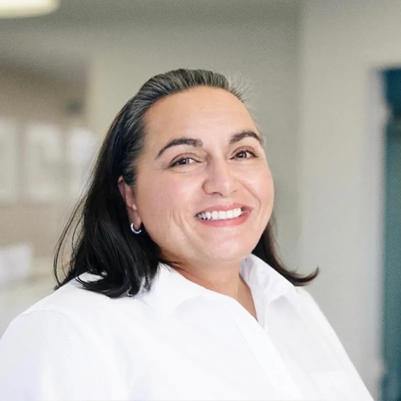 Lächelnde Frau mit dunklen Haaren und weißem Hemd vor unscharfem Hintergrund.