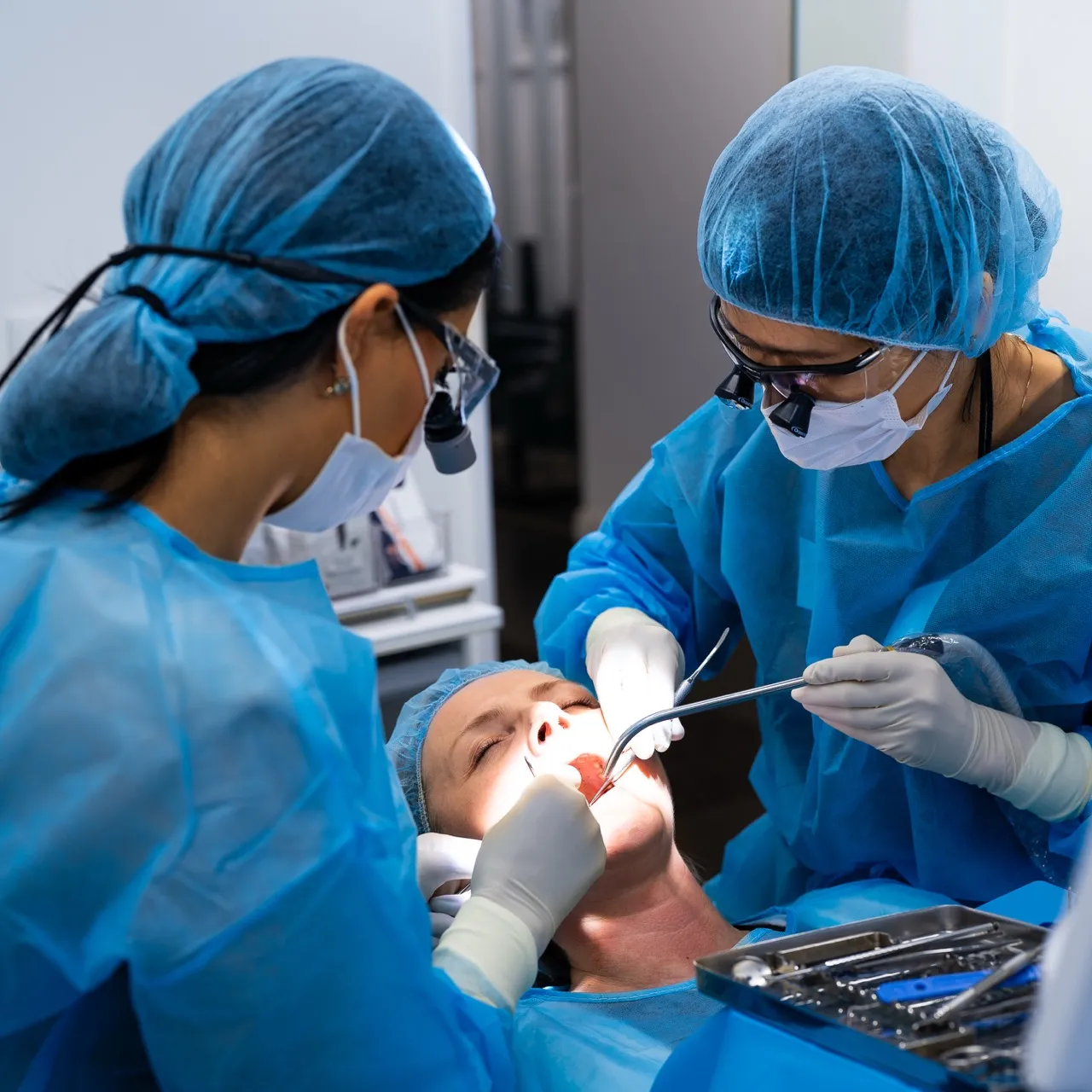 Zwei Zahnärztinnen in blauer Schutzkleidung und OP-Masken behandeln eine liegende Patientin im Mundraum.