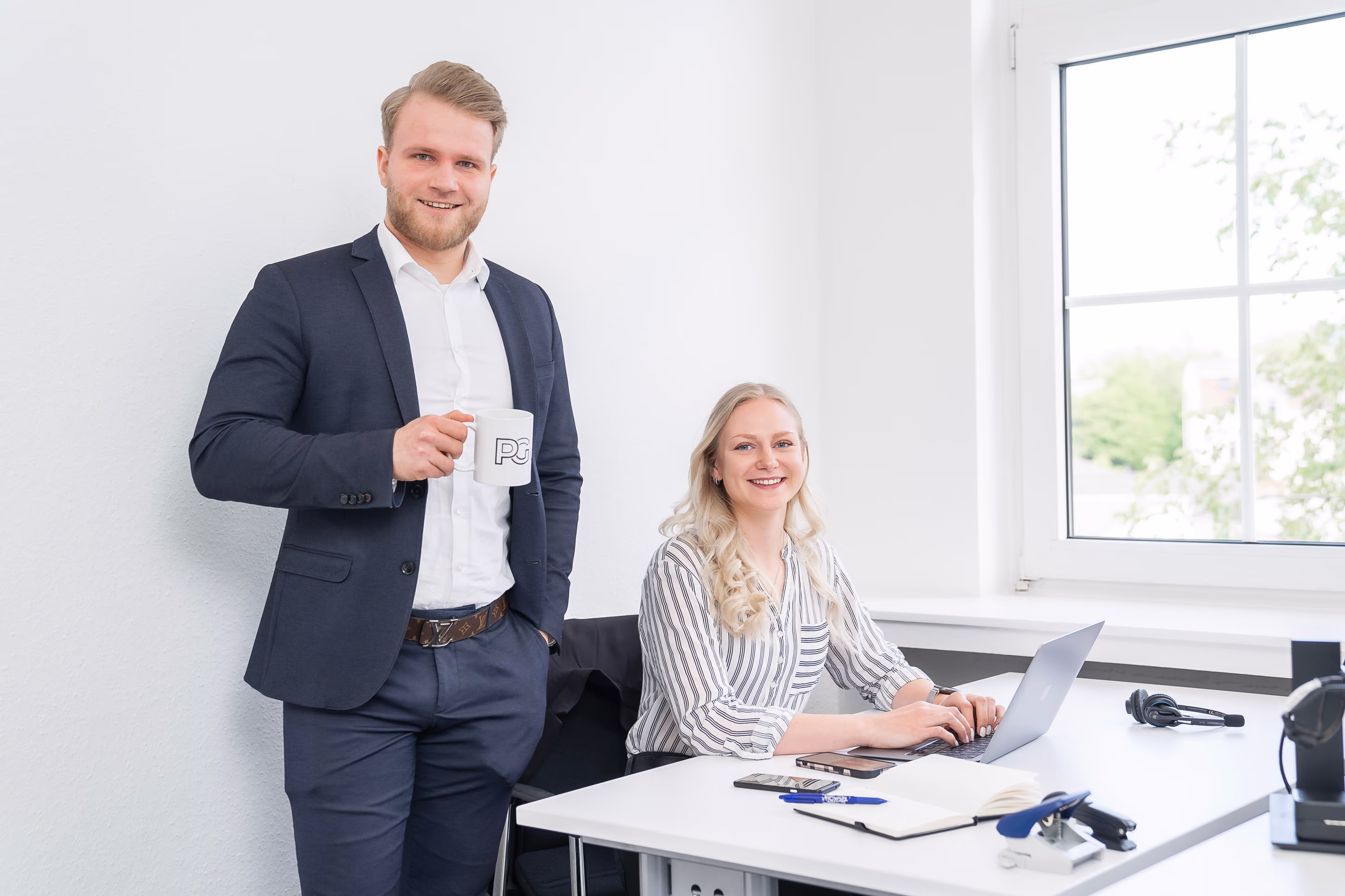 Zwei Teammitglieder im modernen Büro: ein Mann im Anzug mit PG-Tasse in der Hand steht lächelnd neben einer Frau, die am Laptop arbeitet.