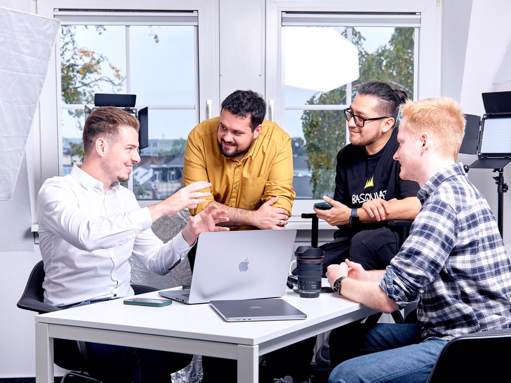 Vier junge Männer in einem modernen Büro bei einem kreativen Teammeeting, umgeben von Kameratechnik und Laptops, lachend und im Gespräch vertieft.
