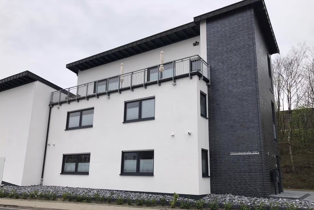 Moderne Außenansicht eines Bürogebäudes mit weißer Fassade, dunklen Fensterrahmen und Balkon im Obergeschoss.