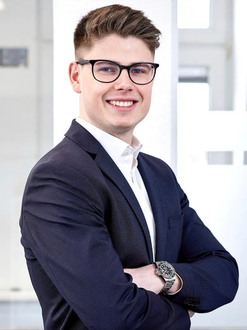 Junger Mann im Anzug mit Brille und Uhr, lächelnd mit verschränkten Armen vor einem hellen Bürohintergrund.