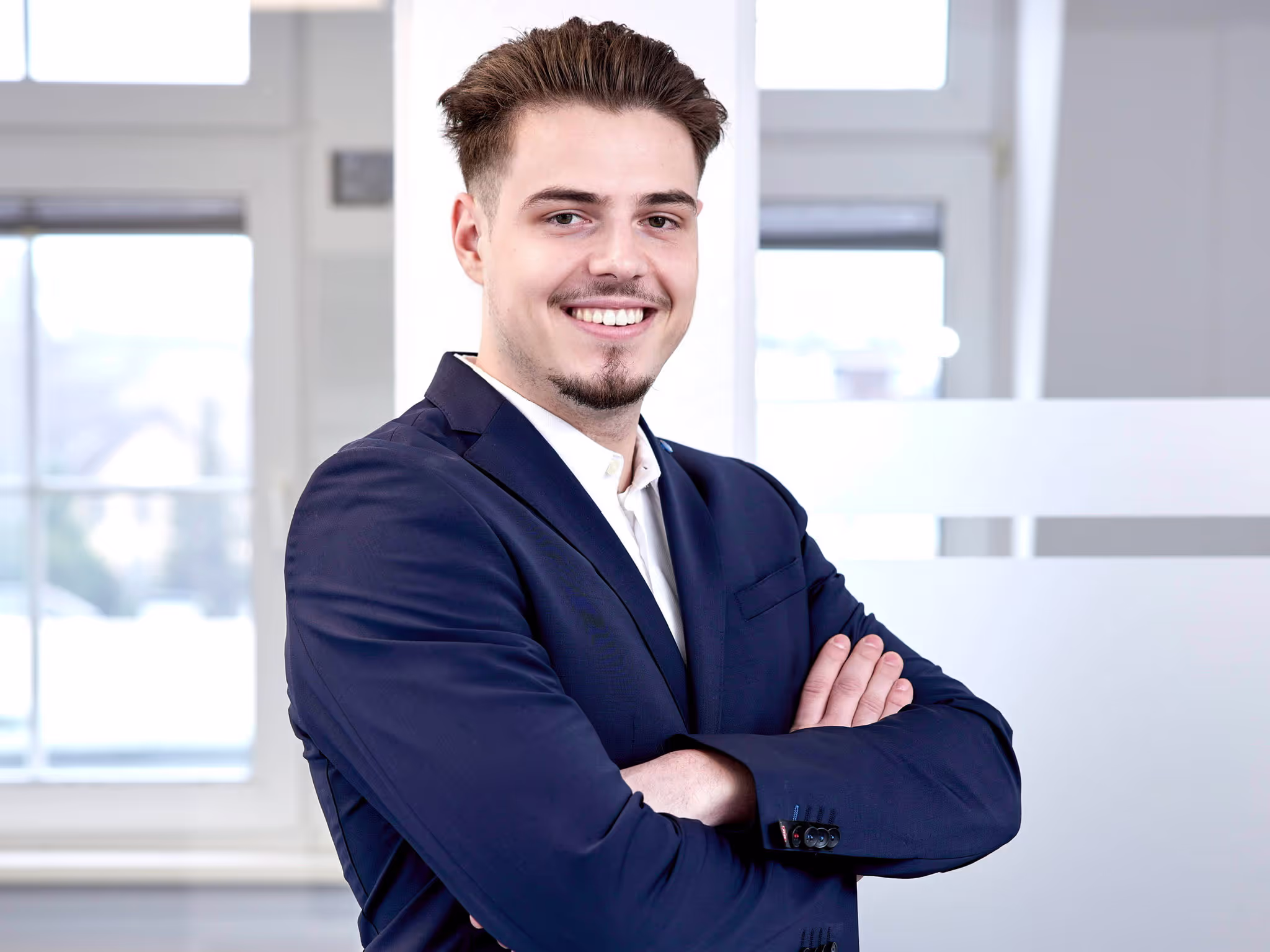 Junger Mitarbeiter im Business-Outfit mit verschränkten Armen lächelt selbstbewusst in die Kamera vor hellem Bürohintergrund.
