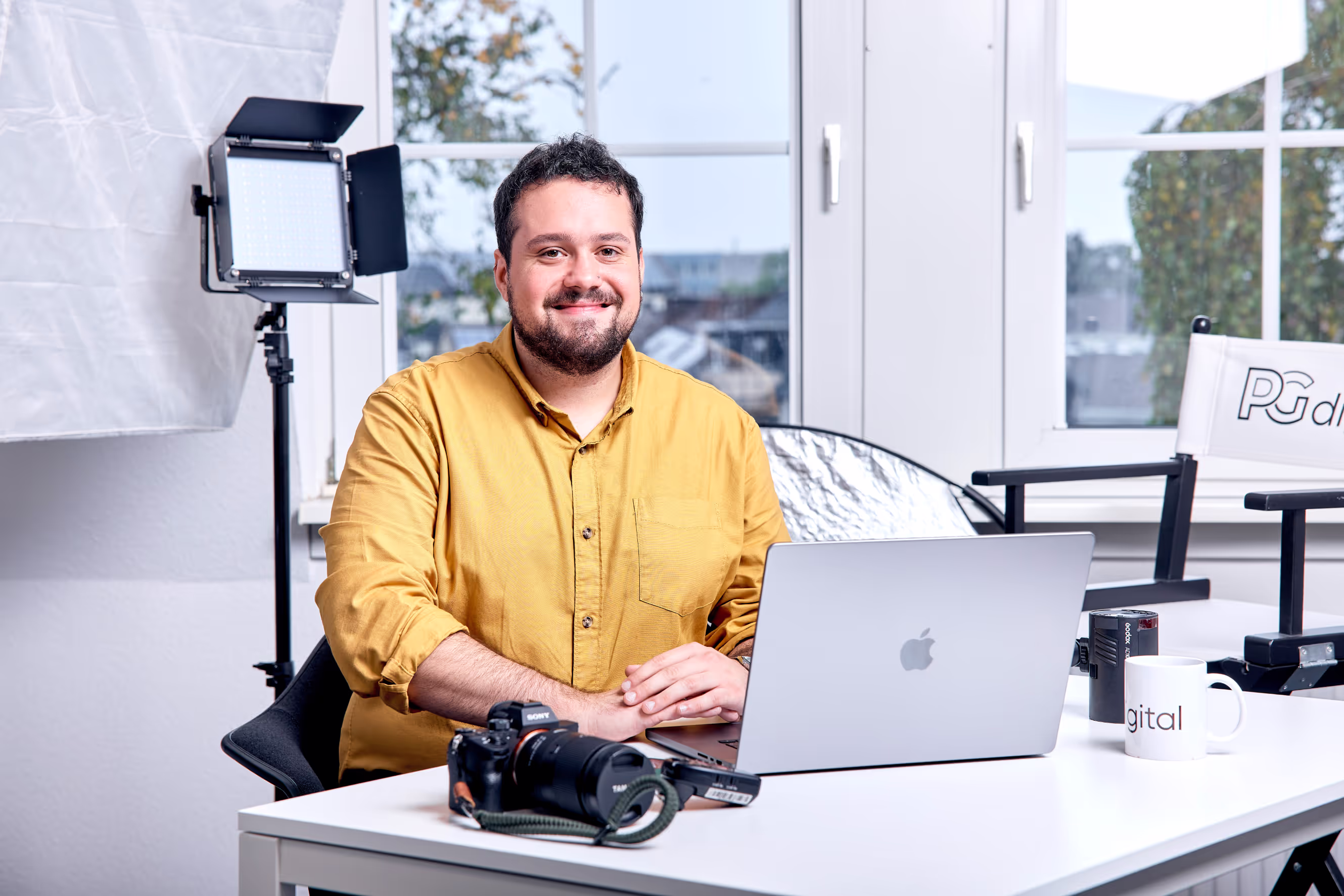 Mann in gelbem Hemd sitzt mit Kamera und Laptop an einem Schreibtisch im Büro von PGdigital, im Hintergrund Studioausstattung.