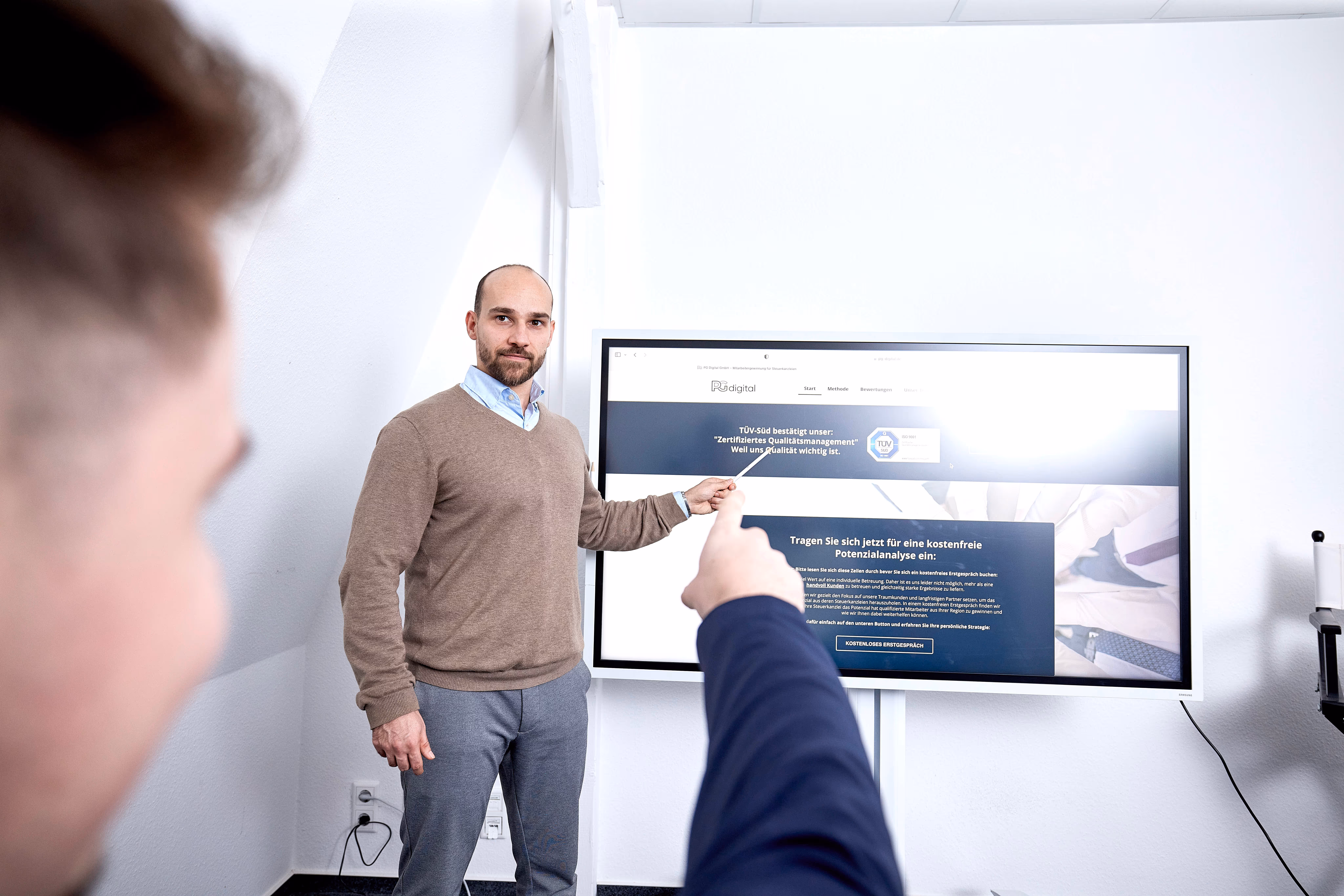 Ein Mann zeigt auf einen Bildschirm mit der Website von PGdigital während einer Präsentation, eine andere Person im Vordergrund deutet ebenfalls auf den Bildschirm.