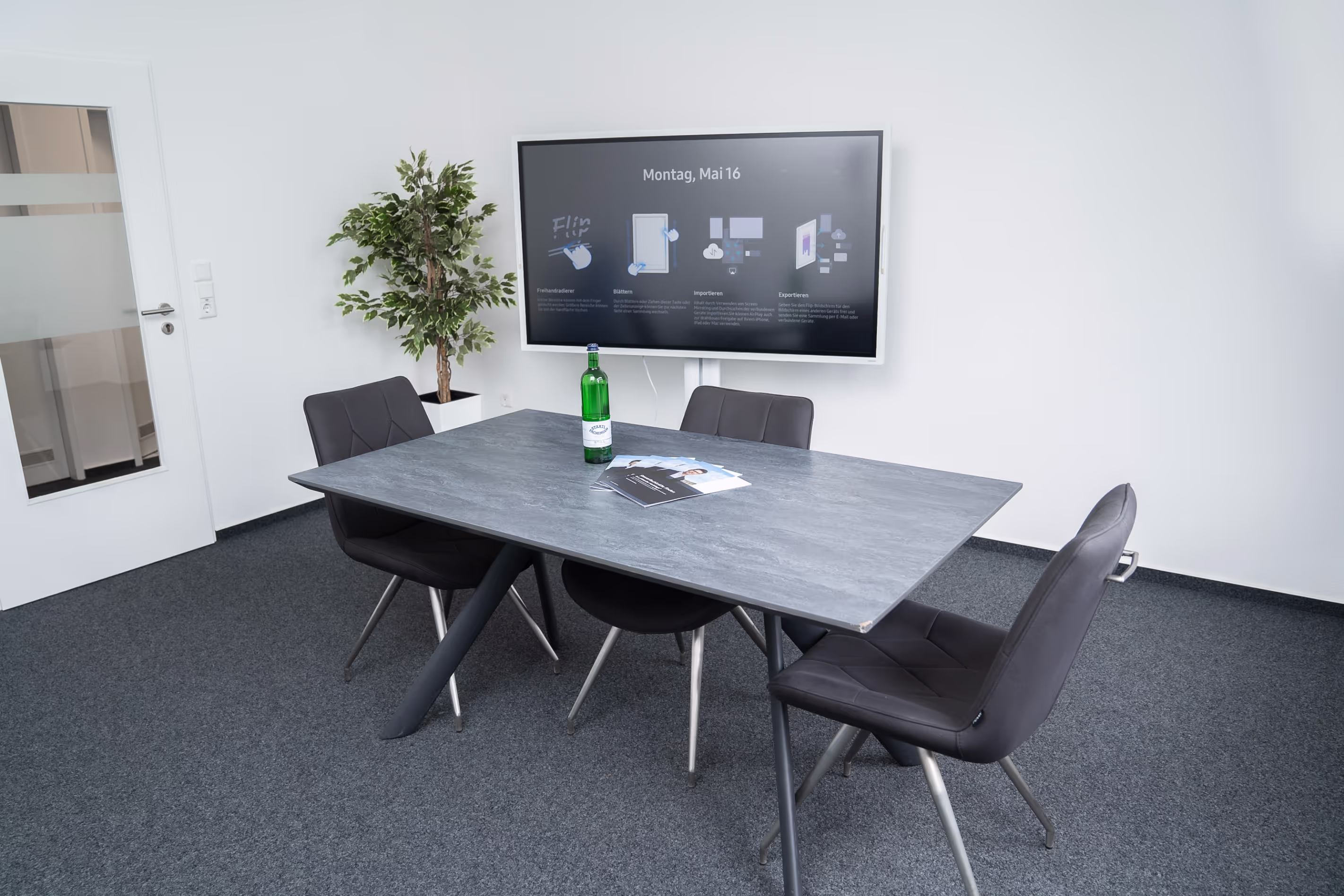Moderner Besprechungsraum bei PG digital mit grauem Tisch, vier Stühlen, Wandbildschirm und Deko-Pflanze – bereit für Meetings oder Präsentationen.