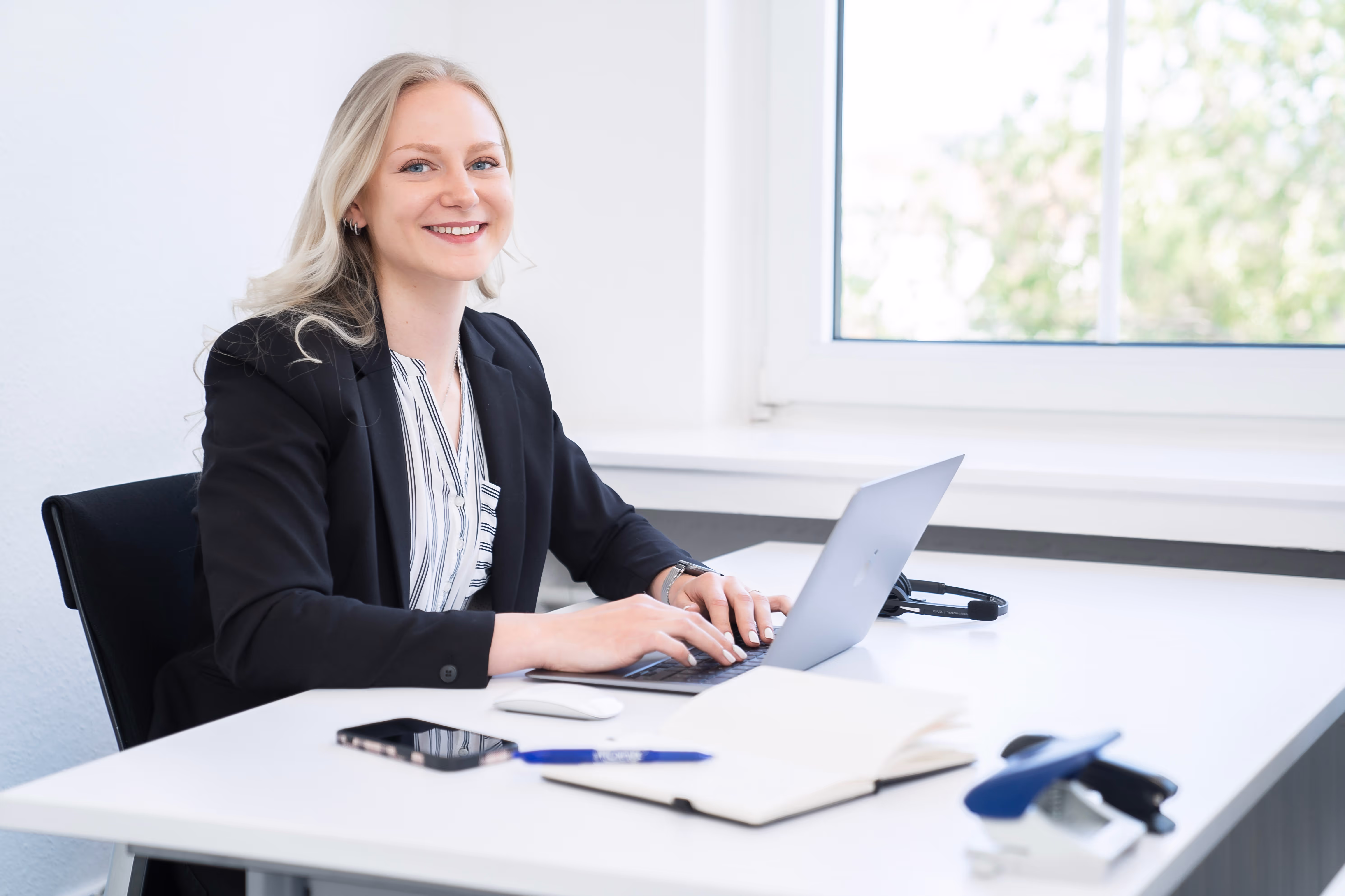 Eine lächelnde Mitarbeiterin im Business-Outfit sitzt an einem weißen Schreibtisch und arbeitet an einem Laptop. Auf dem Tisch liegen ein Notizbuch, ein Stift, ein Smartphone sowie ein Headset – im Hintergrund ein helles Büro mit großem Fenster.
