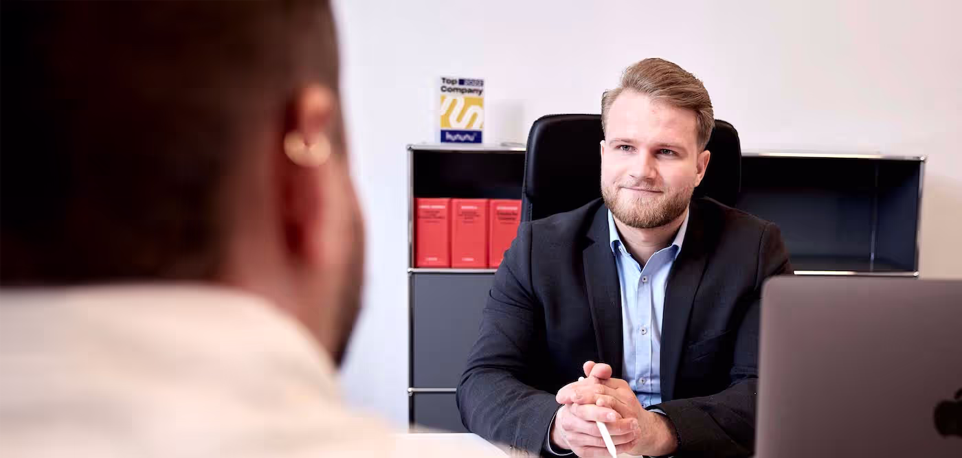 Ein Mann im Anzug führt ein Beratungsgespräch in einem modernen Büro. Er sitzt hinter einem Schreibtisch, hört aufmerksam zu und schaut seinen Gesprächspartner an. Im Hintergrund stehen rote Gesetzestexte und ein Kununu-Siegel „Top Company“.