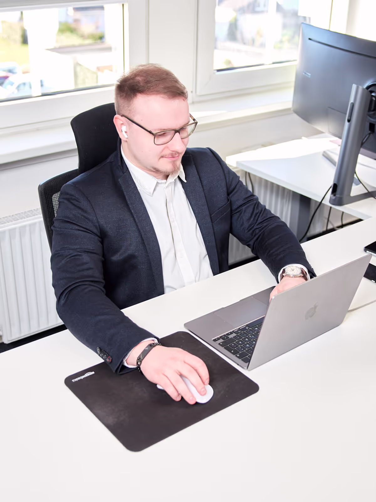 Ein junger Mann mit Brille und AirPods arbeitet konzentriert an einem Laptop in einem modernen Büro. Er trägt ein dunkles Sakko und nutzt eine externe Maus auf einem schwarzen Mauspad, im Hintergrund steht ein großer Monitor.