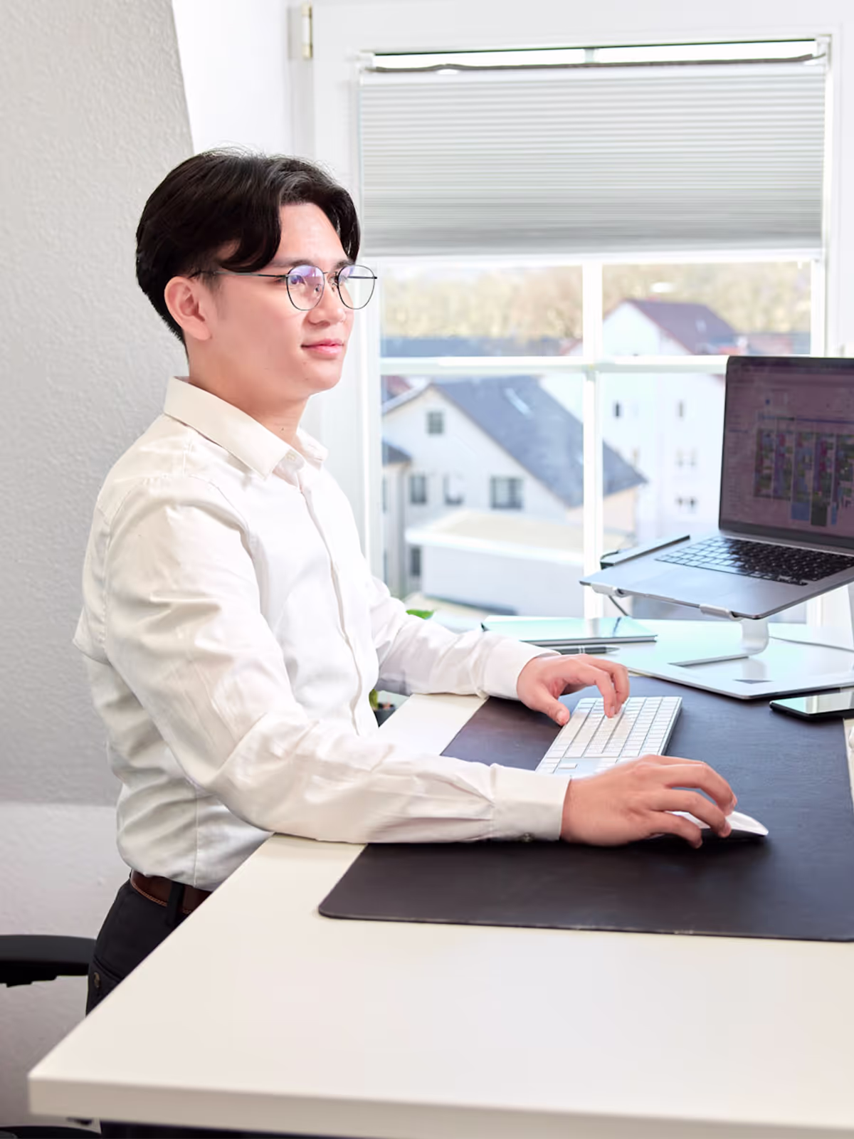 Ein junger Mann mit Brille arbeitet fokussiert an einem höhenverstellbaren Schreibtisch mit Laptop und externer Tastatur. Im Hintergrund sind große Fenster mit Blick auf Dächer und Bäume zu sehen.