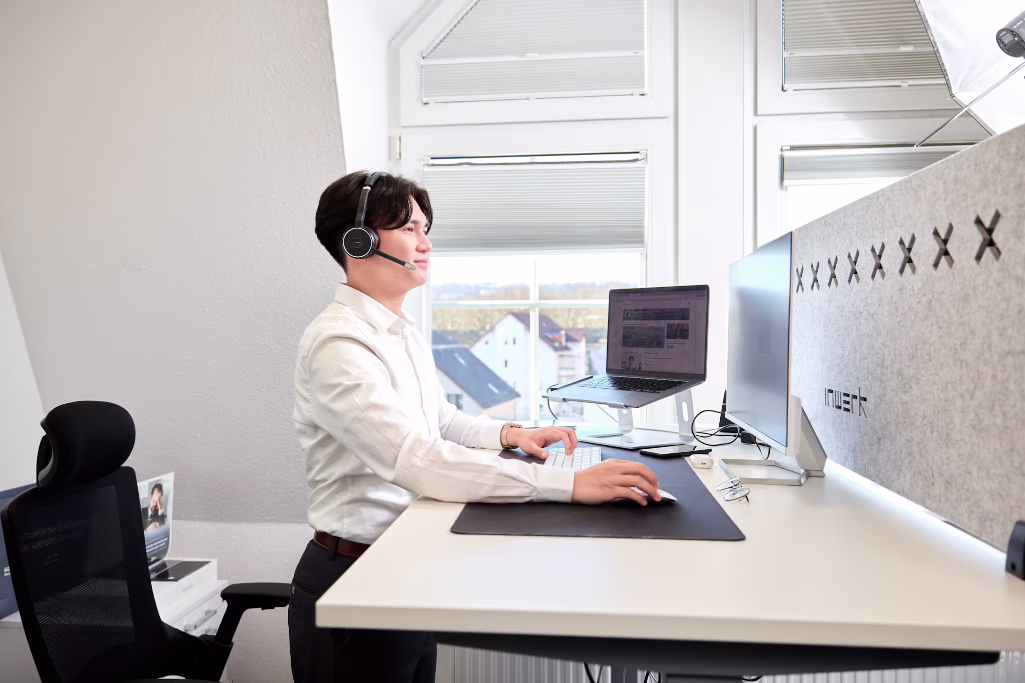 Ein junger Mitarbeiter mit Headset steht an einem höhenverstellbaren Schreibtisch und führt einen Videocall über seinen Laptop. Der Arbeitsplatz ist modern eingerichtet mit zwei Monitoren und Blick aus dem Fenster.