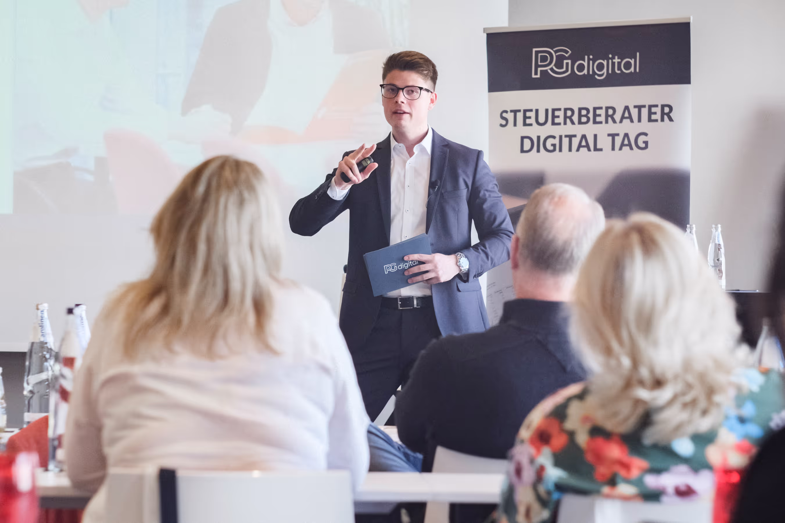 Ein Redner im Anzug präsentiert beim Steuerberater Digital Tag vor einem Publikum. Im Hintergrund ist ein PGdigital-Roll-Up sichtbar, im Vordergrund sitzen Teilnehmende und hören aufmerksam zu.
