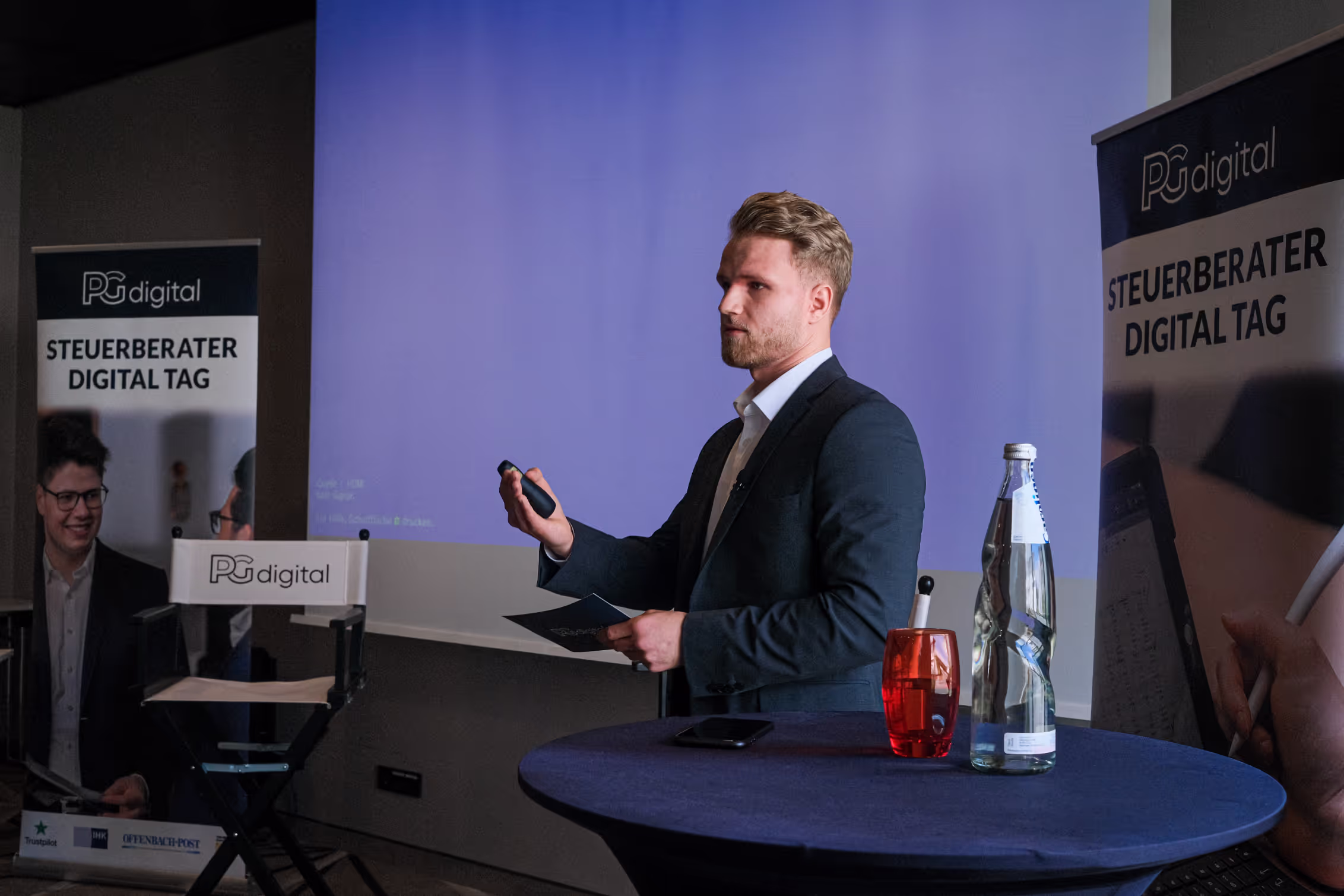 Ein Redner hält eine Präsentation beim Steuerberater Digital Tag von PGdigital. Im Hintergrund stehen zwei Roll-Ups mit dem Veranstaltungslogo, eine Leinwand mit Präsentation sowie ein Tisch mit Getränken und Mikrofon.
