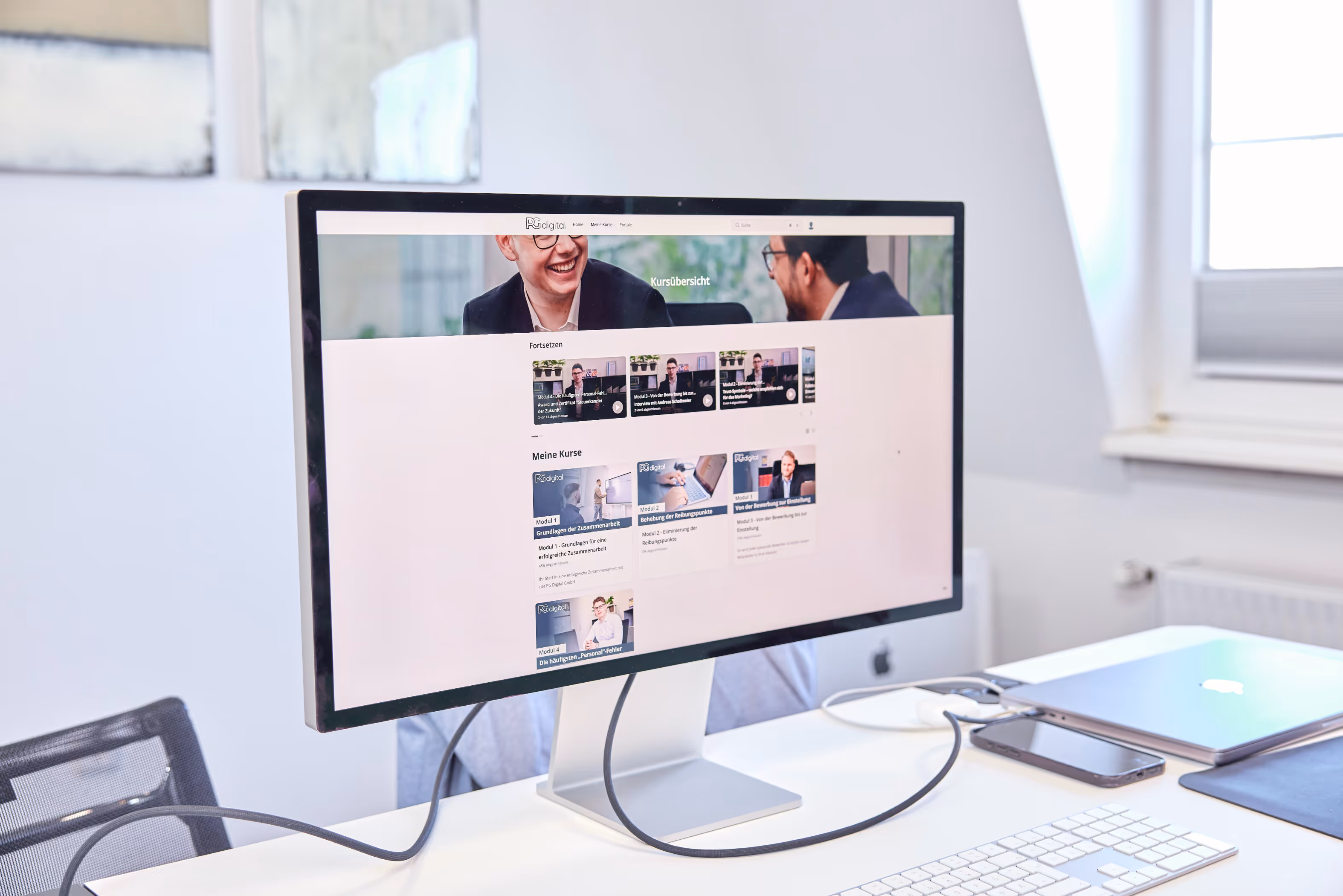 Ein moderner Arbeitsplatz mit einem großen Monitor, auf dem eine Online-Lernplattform mit mehreren Kursen angezeigt wird. Daneben liegen ein MacBook, ein Smartphone und eine Tastatur auf einem weißen Schreibtisch.
