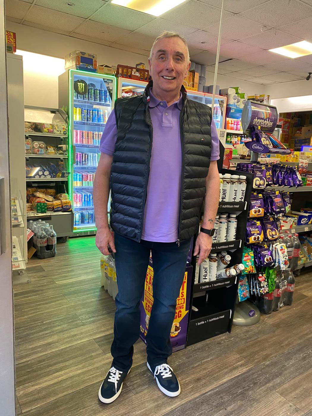 Smiling man wearing a black puffer vest, purple polo shirt, blue jeans, and sneakers standing inside a convenience store.