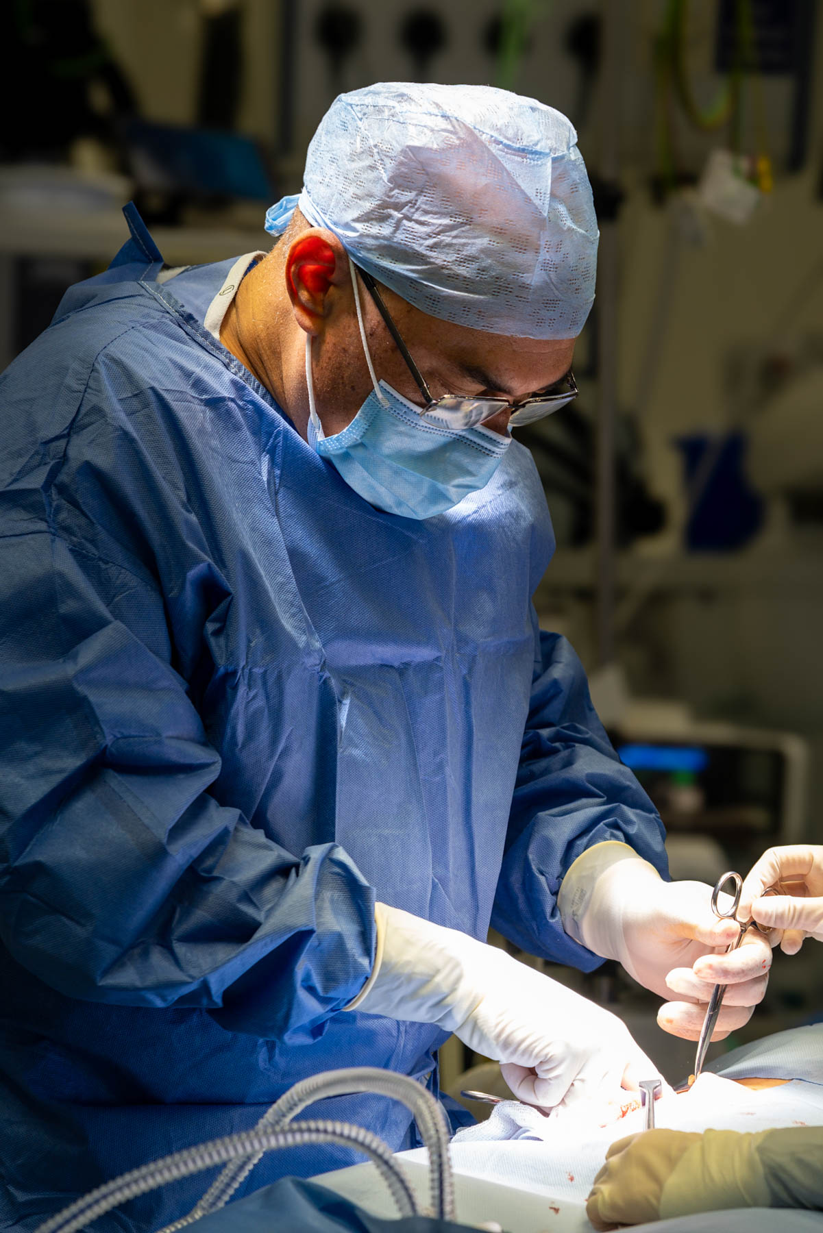 Surgeon wearing blue scrubs, surgical cap, mask, and gloves performing an operation in a well-lit operating room.