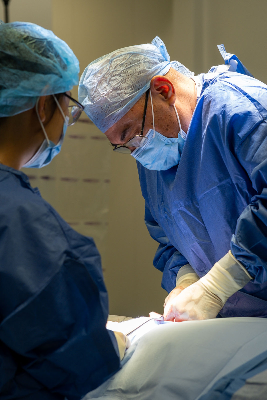 Two surgeons in blue scrubs and surgical masks performing surgery under bright lights.