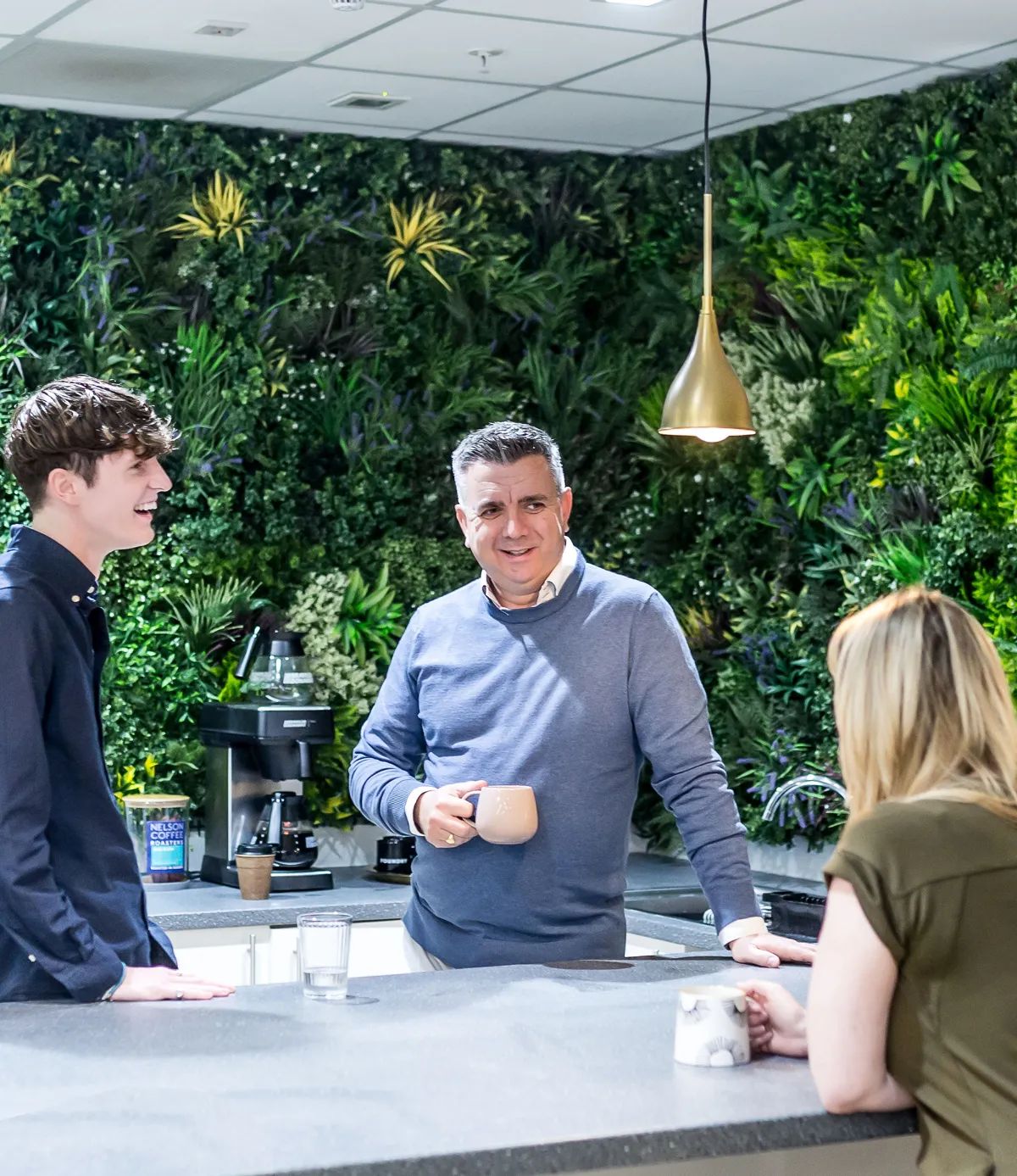 Consultant with a mug in hand talking and laughing with clients in an office kitchen with greenery.