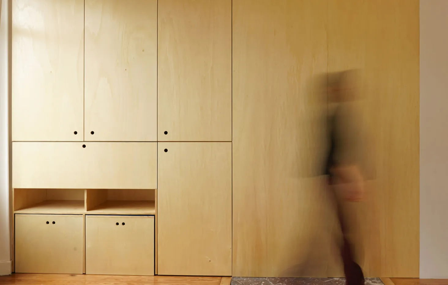 Personne floue en mouvement devant un mur et des armoires en bois clair avec des poignées noires.