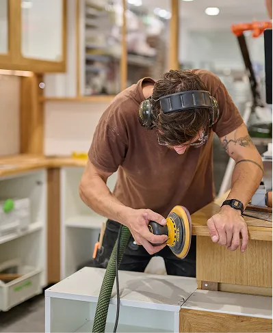 Homme portant un casque antibruit et utilisant une ponceuse électrique pour lisser un meuble en bois dans un atelier.