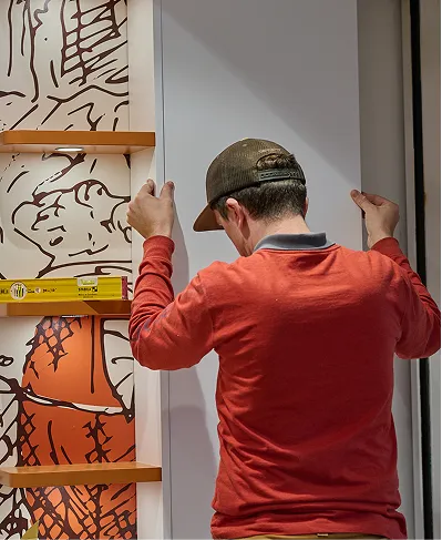 Homme en casquette et sweat rouge installant ou ajustant une porte blanche à côté d'une étagère orange.