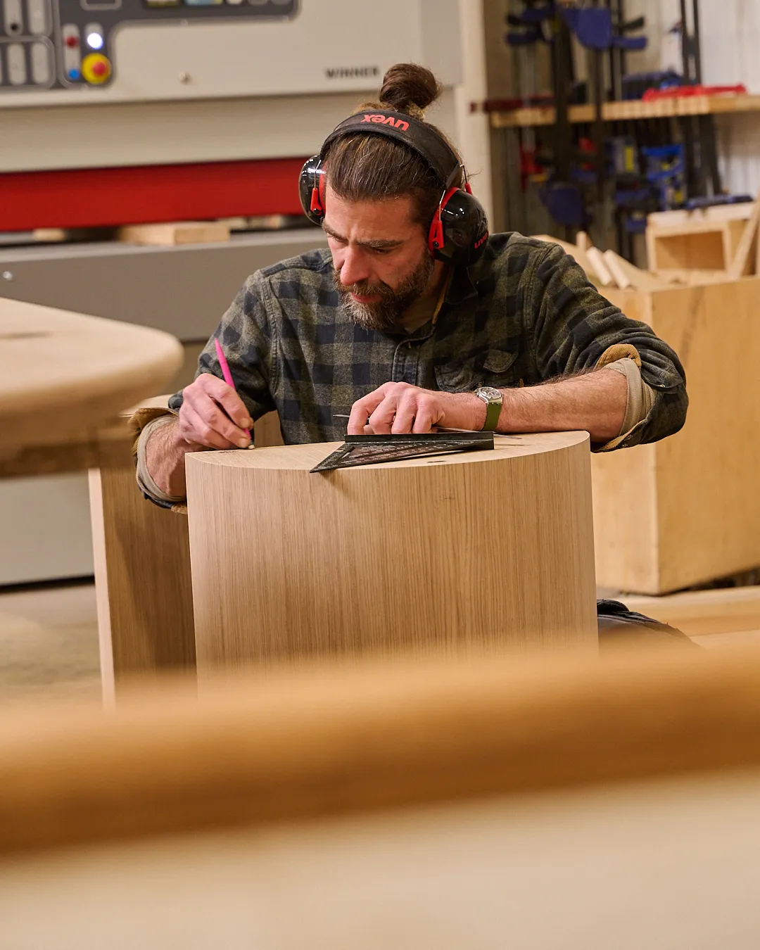 Un homme avec une chemise à carreaux et des protections auditives trace des mesures sur un meuble en bois clair dans un atelier de menuiserie.
