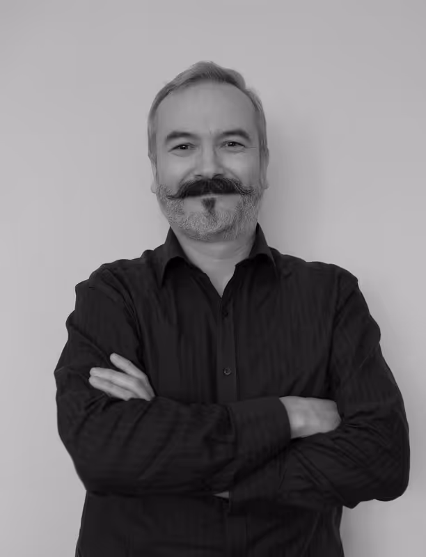 Smiling middle-aged man with a mustache and beard, wearing a black shirt with arms crossed against a plain background.