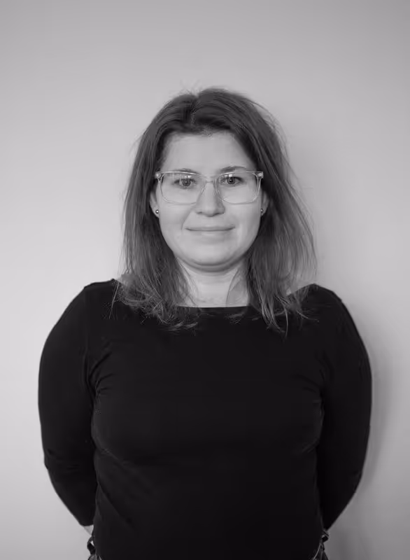 Smiling woman with shoulder-length hair wearing clear glasses and a black top against a plain background.