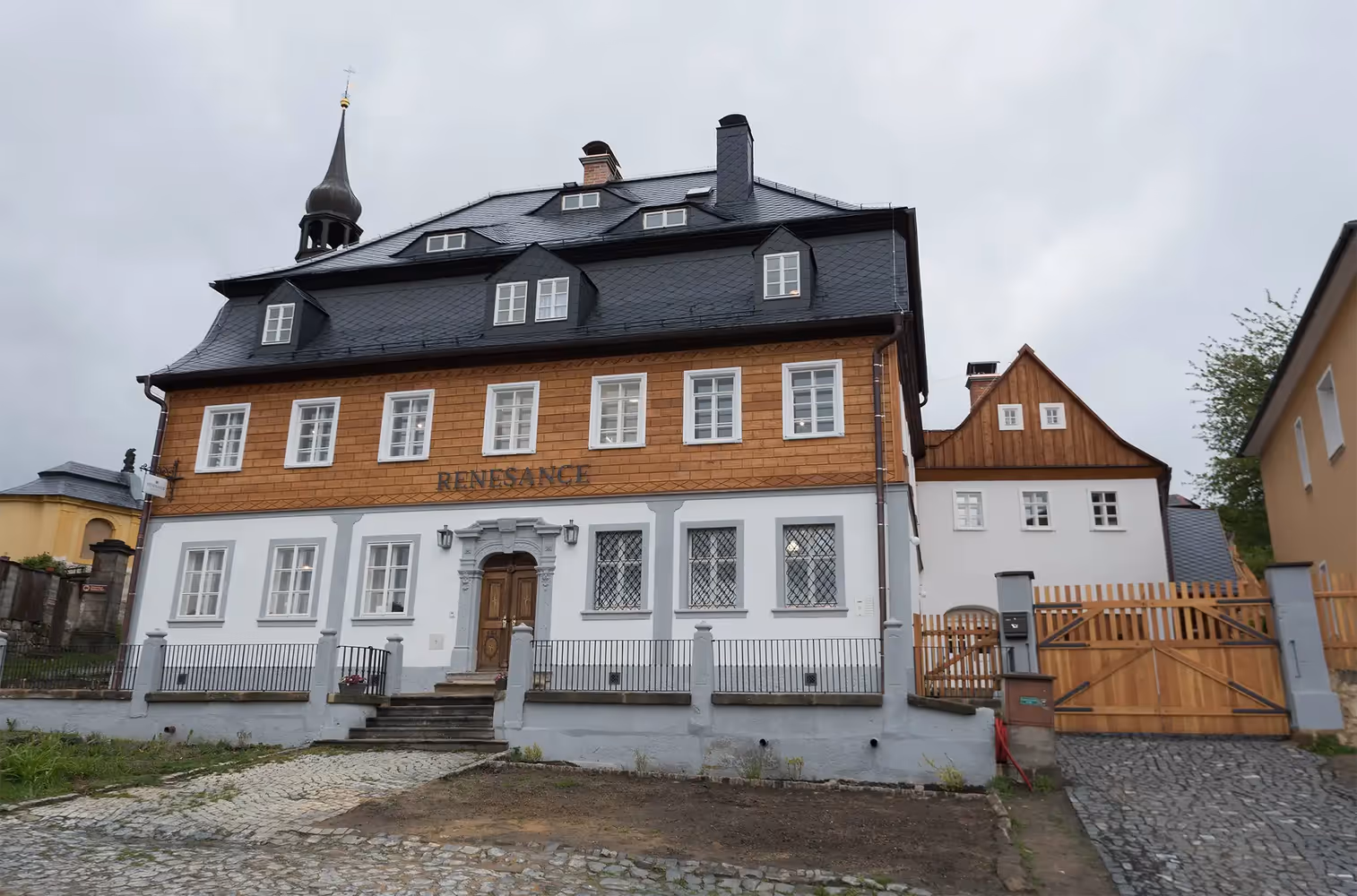 Historic two-story building with a black steep roof, wooden upper facade, white lower facade, and a central wooden door under a decorative stone arch.